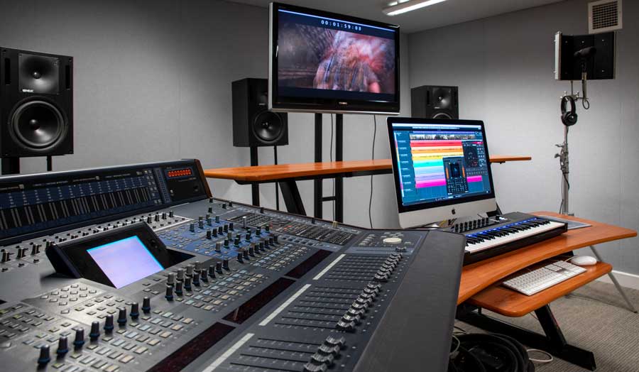 a large mixing deck at the front of the photo, 2 screens and 2 keyboards further away and 3 large speakers in the background