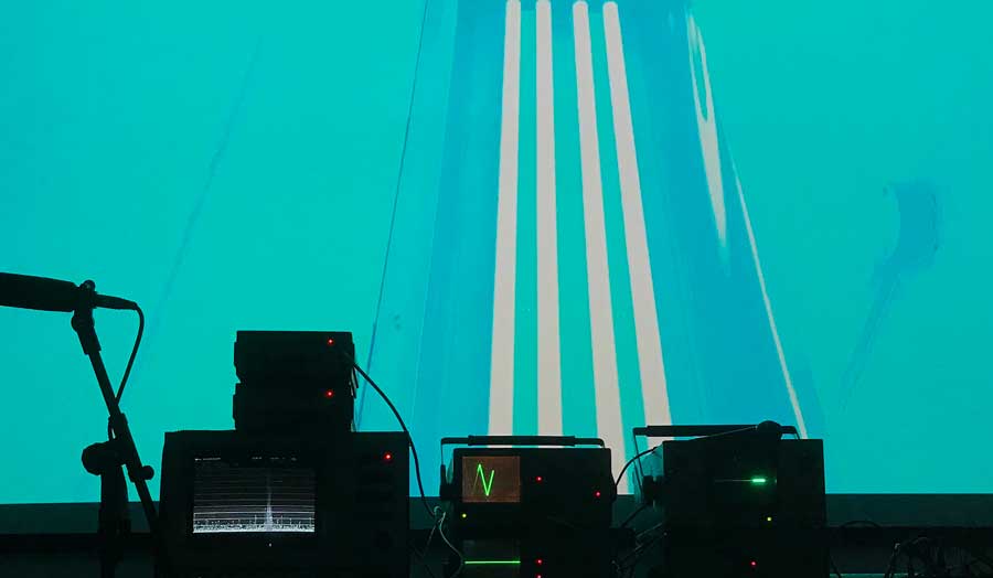 3 pieces of equipment and a standing microphone in the dark in the forefront, in front of them a wall lit up in blue with white stripes in the middle