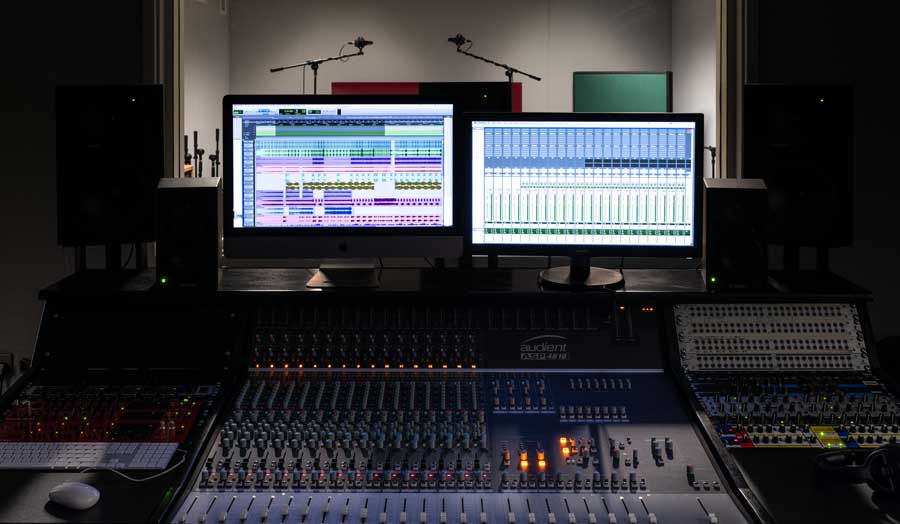 a mixing deck and 2 screens in a music control room, a live recording room behind them