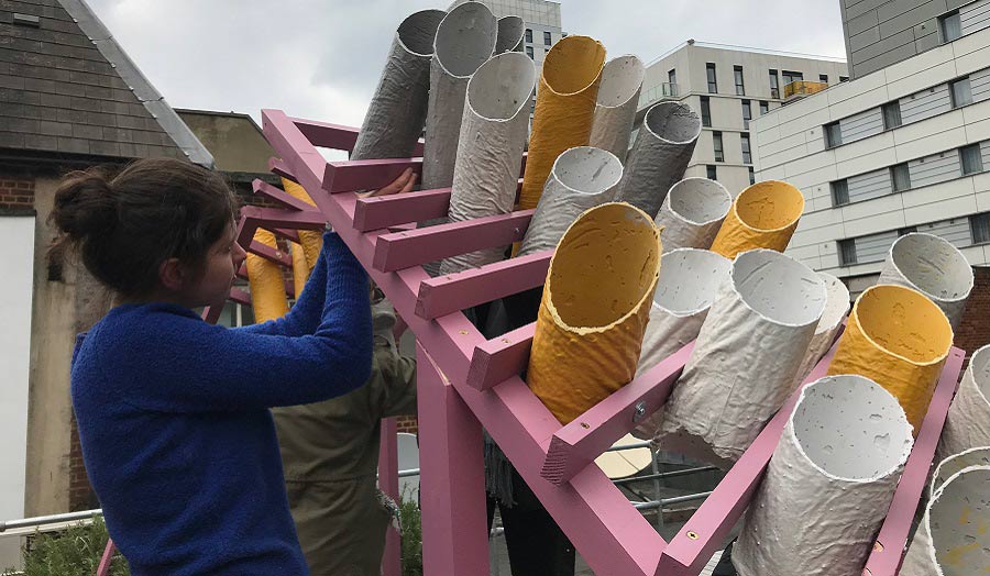 A sculpture on the rooftop of a building made of cardboard painting tubes going through a pink wooden structure