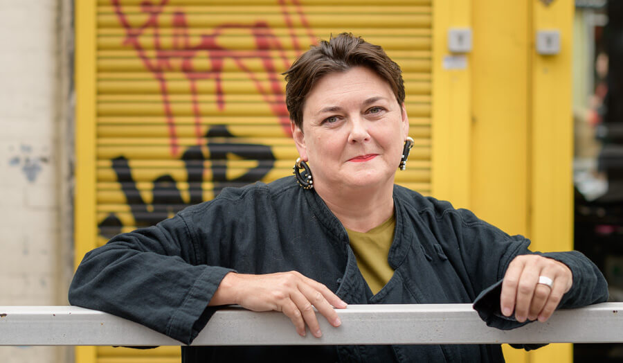 Short haired female lecturer Harriet McKay smiles to the camera as she stands against some graffiti.