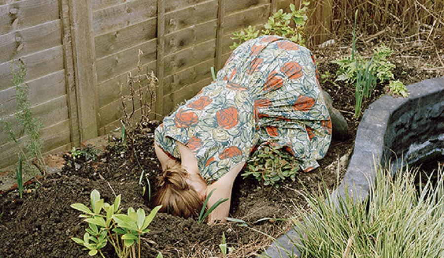 Woman kneels in garden with head buried in hole in soil