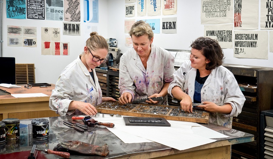 Three women look at traditional letter press in a print-room