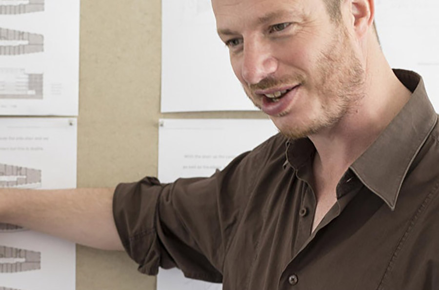 A close up portrait of a man who is talking and with his arm outstretched pointing to some diagrams pinned to the wall