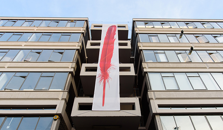 Photograph of a canvas with a red feather attached to the outside of The Cass building.