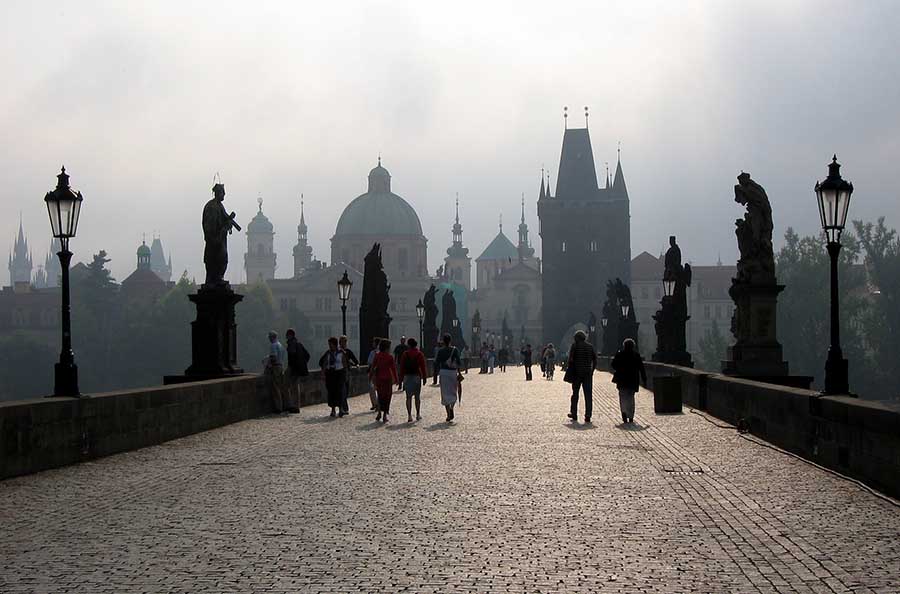 A view over a wide pedestrian bridge with a silhouette of Prague beyond.