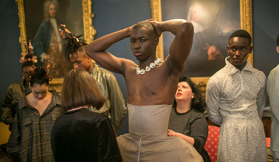 Male model being fitted with a corset in a gallery surrounded by historic paintings