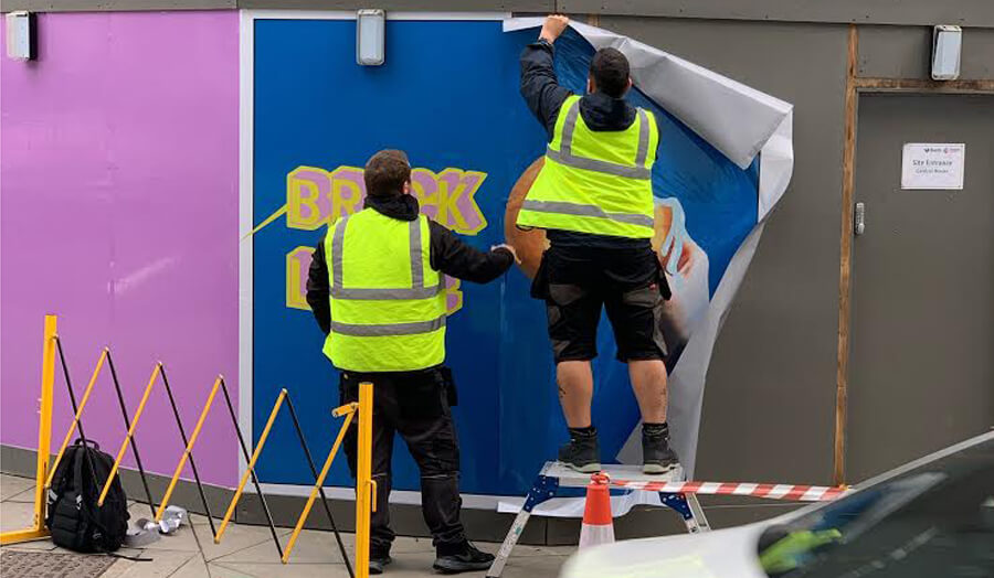 2 men in hi-vis jackets putting up a blue poster on building hoardings