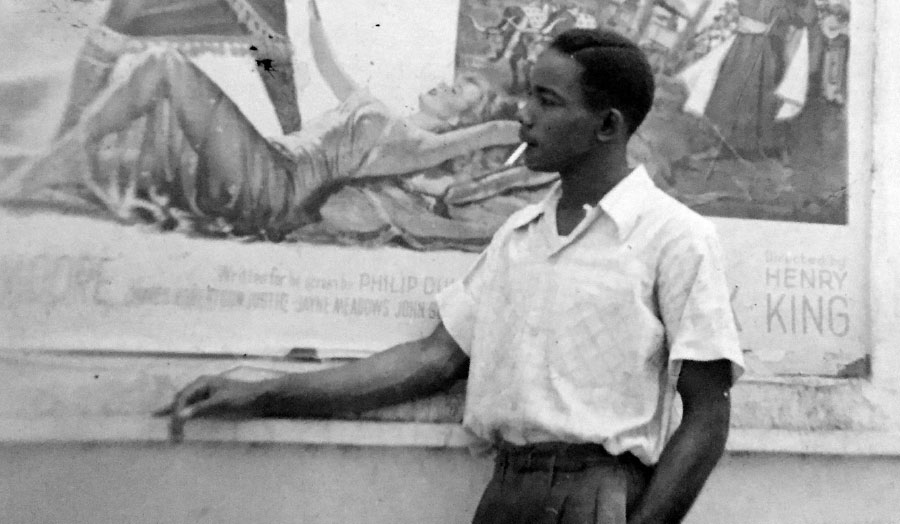 Unknown man with cigarette, white shirt, in front of a poster, Kingston Jamaica, circa 1950
