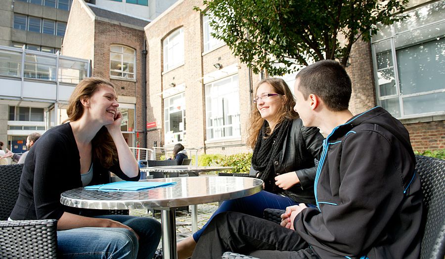 three students sat at a table outside in the courtyard at holloway talking and smiling 