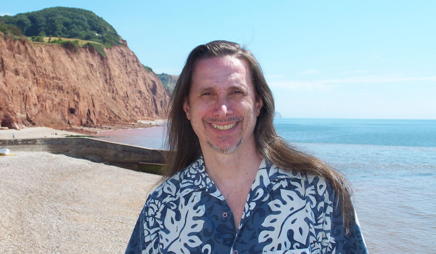 Long-haired male lecturer Manfrendo Meraviglia stands on a beach on a sunny day smiling to camera.