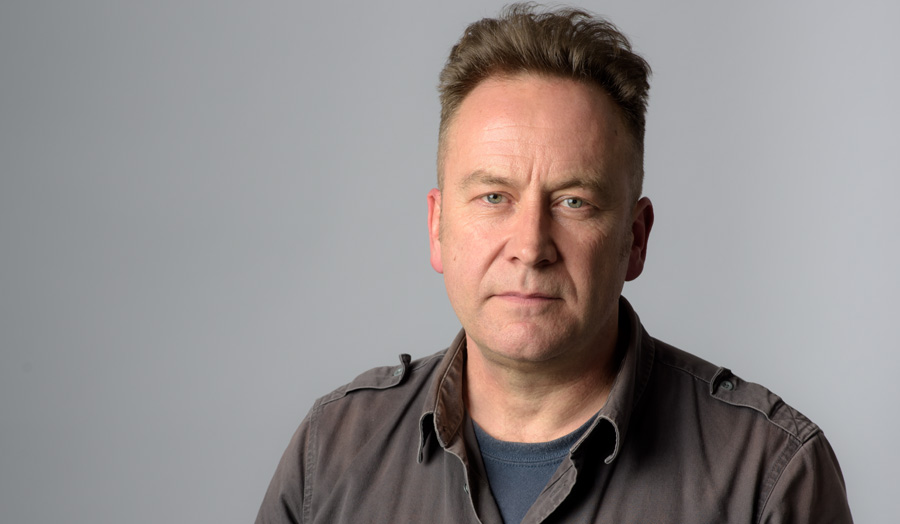 Simon Cadman, wearing a brown shirt, faces the camera directly against a grey background.