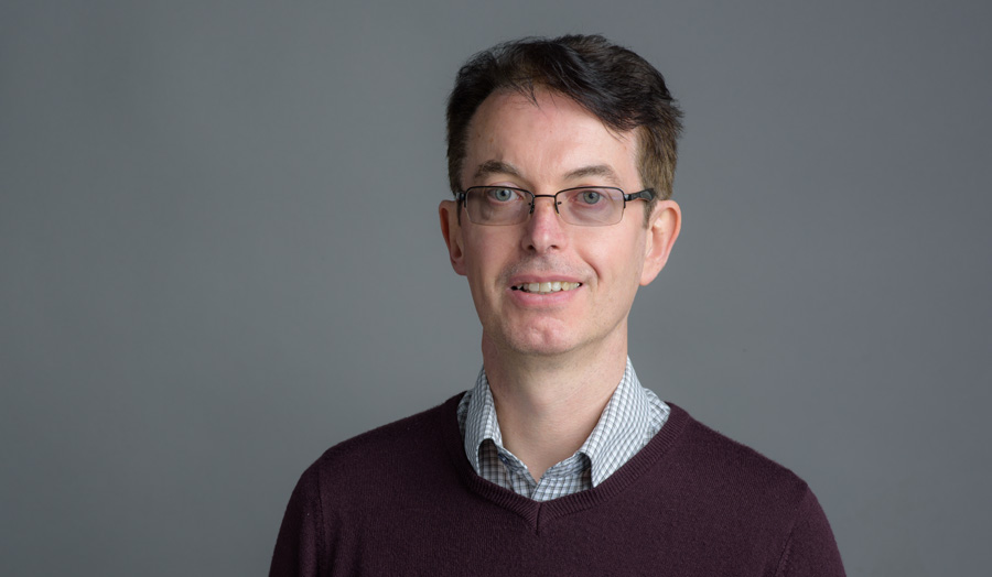 Bespectacled male lecturer Stephen Breen smiles to the camera against a grey background.