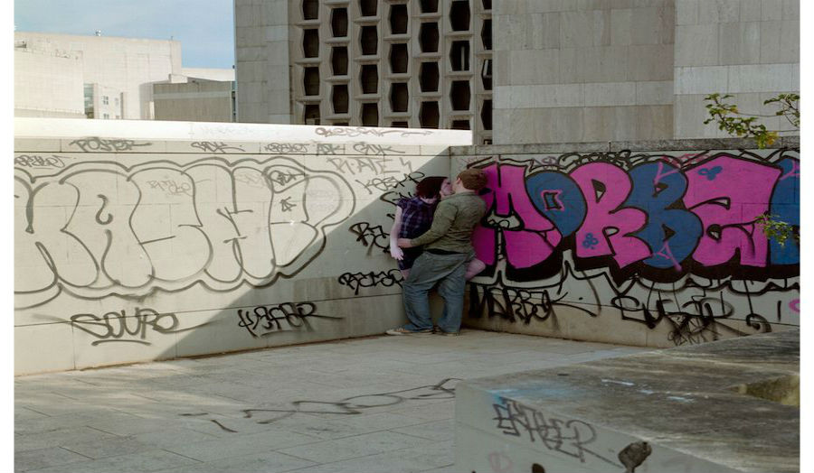 A young couple kissing in the corner of graffiti-riddled skate park. 