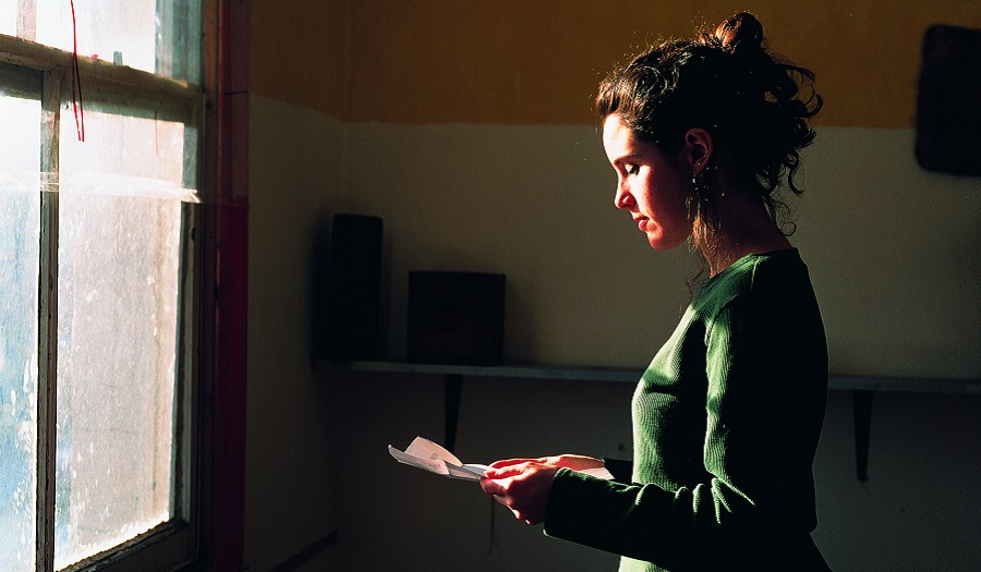 A woman standing in front of a window looks down at a letter.