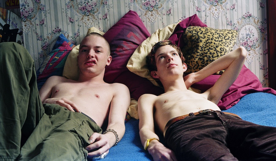 two topless young men recline on a bed looking towards viewer