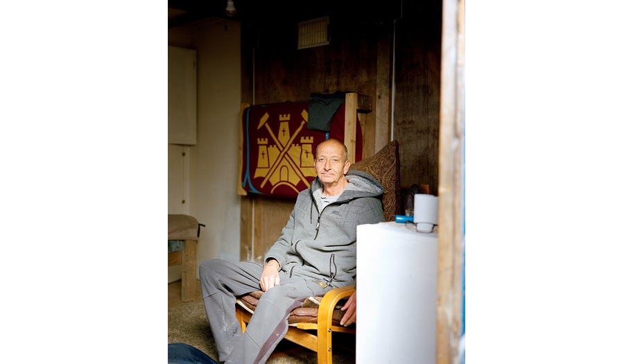 An elderly man sits in a shed, looking out towards the camera