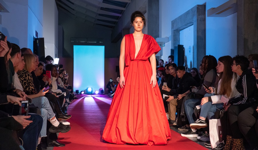 a woman in a long red dress stand on a fashion catwalk with n audience seated on either side
