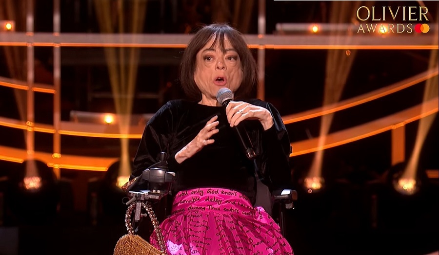 A woman in a wheelchair speaks into a microphone with a black and gold background