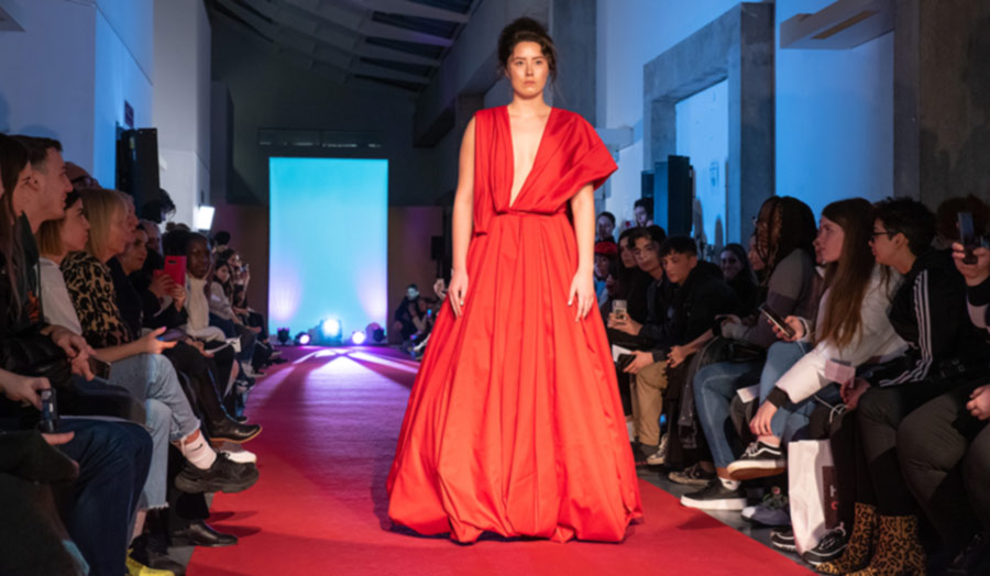A female model in a long red dress walking through a fashion show with audience members sitting on either side of the catwalk