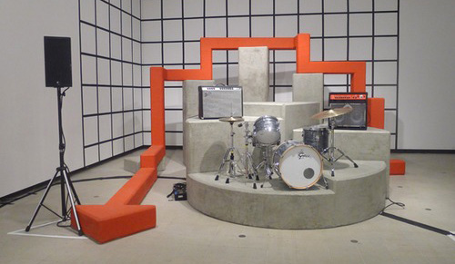 A drum kit is set up on concrete plinths. A large orange arrow sculpture wraps around the room.
