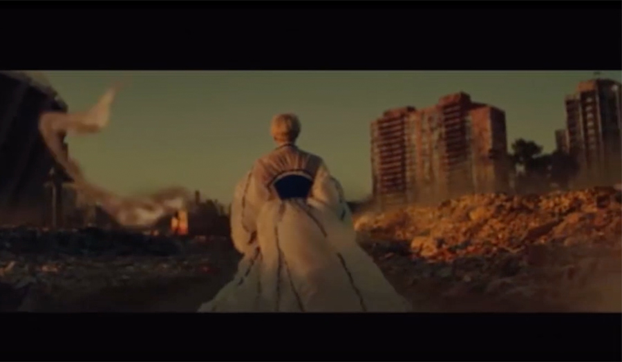 A still image from a film, showing the back of a figure wearing a dress. Some housing blocks are visible in the distance.