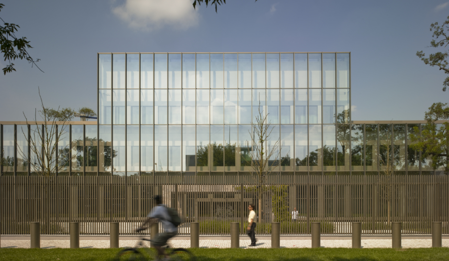 Colour photo of gridded glass facade above wooden structure with trees, cyclist and pedestrian