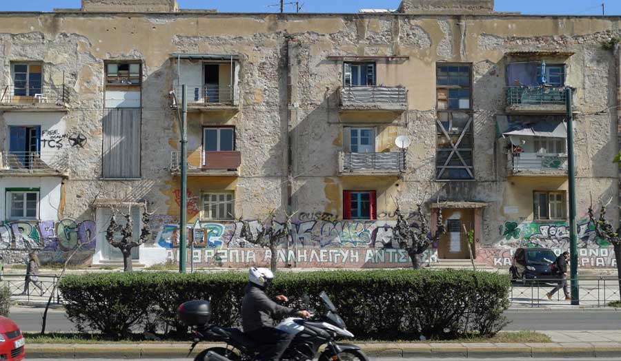 Elevational view of a run-down housing block with balconies, with a motorcyclist passing in front

