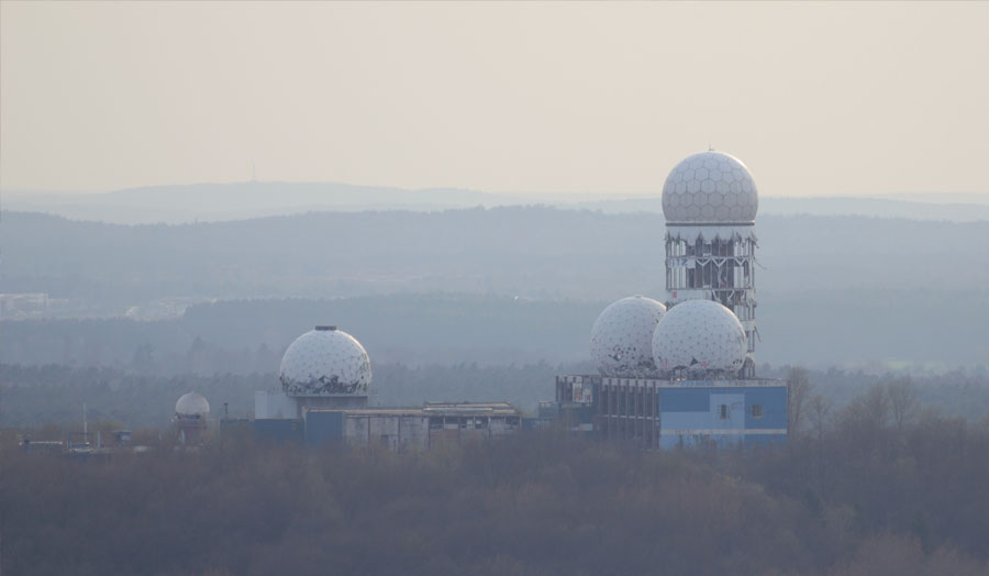 A misty view across the tops of trees to further forested hills on the horizon. In the middle distance a series of large structures rise from amongst the trees. Their surfaces are decayed and their windows are broken. The structures include wide buildings of 1 to 3 storeys and a tall tower, they are topped by large white domes which are also broken and decayed. 