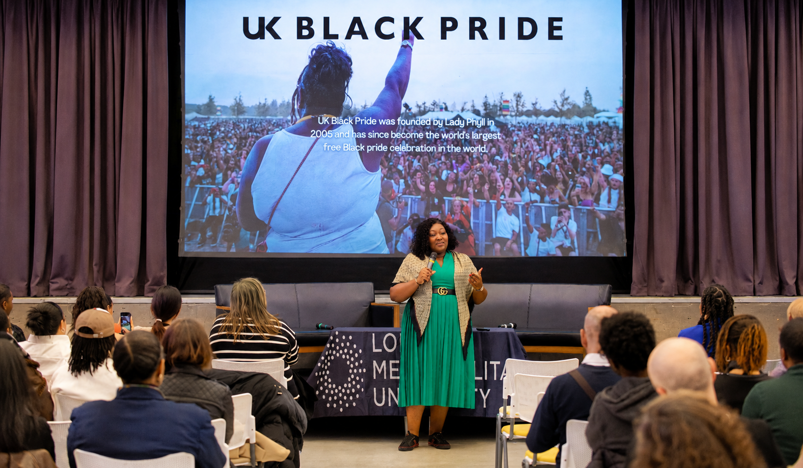 A speaker addresses an audience during a UK Black Pride event, standing in front of a large screen
