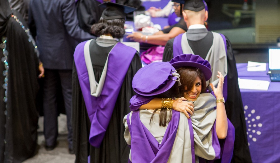 two female graduates in full robes embrace during graduation with one smiling at camera 
