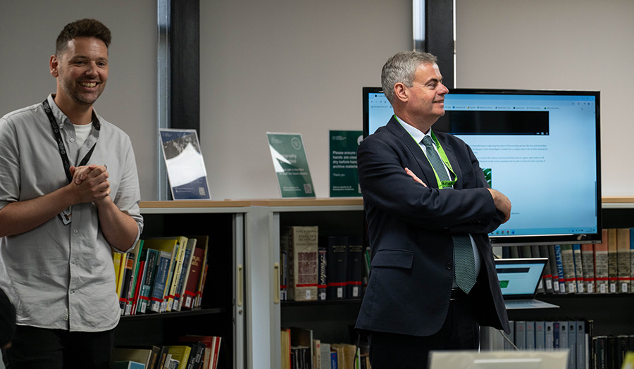 Two men standing in a library, one wears a suit and green tie and one wears a white shirt