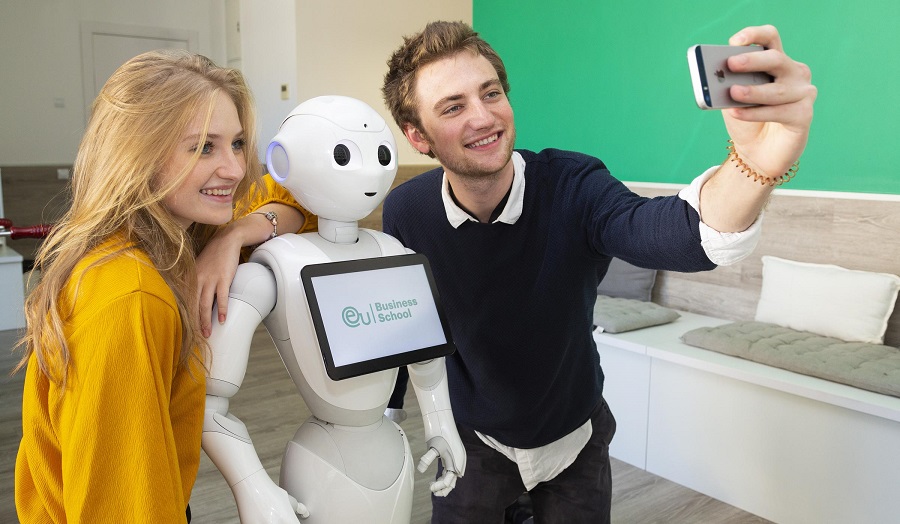 a young man and a young woman taking a selfie with a robot with an 'EU business school' logo on it