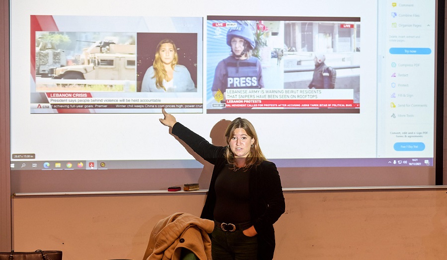 Woman presenting a lecture, pointing to a newspaper heading on a screen