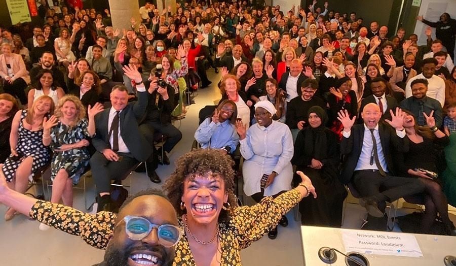 Two hosts of an award ceremony, taking a selfie with the audience in the background