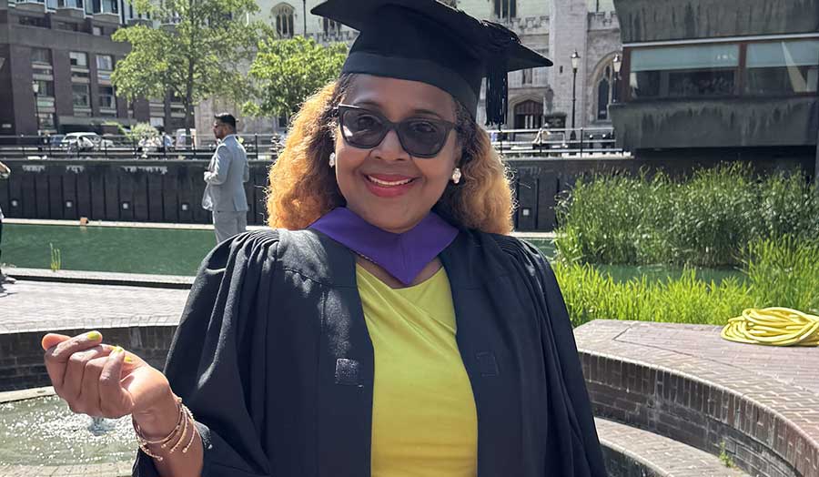 A woman in a yellow dress wearing a cap and gown smiles at graduation