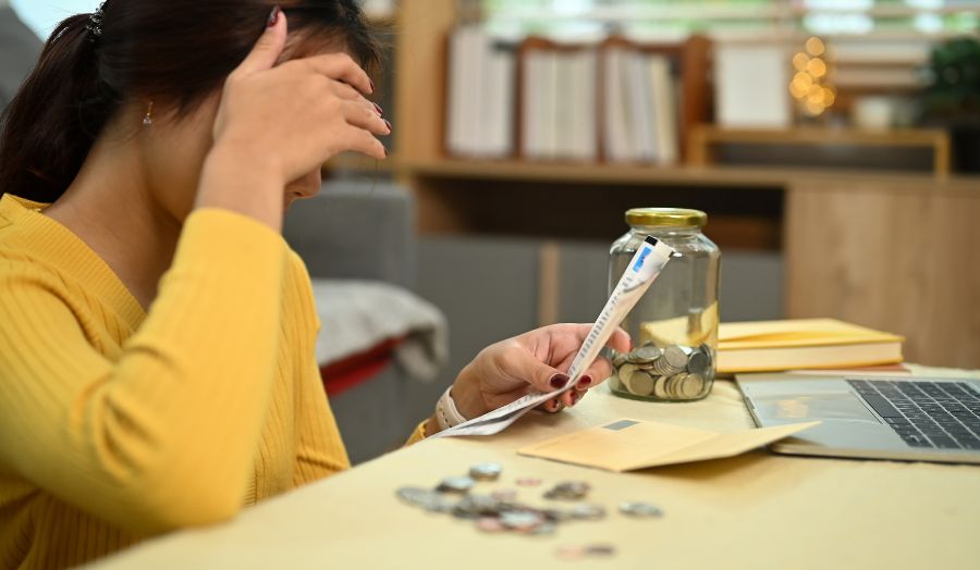 A woman sits at a table reading bills, there is a jar with coins on the table