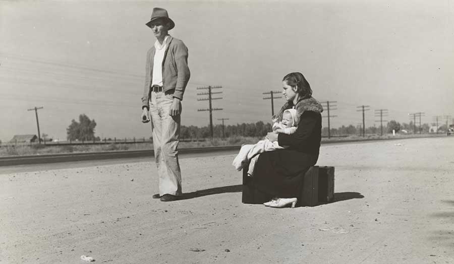 Man in a hat and women holding a child and sitting on the suitcase on the road