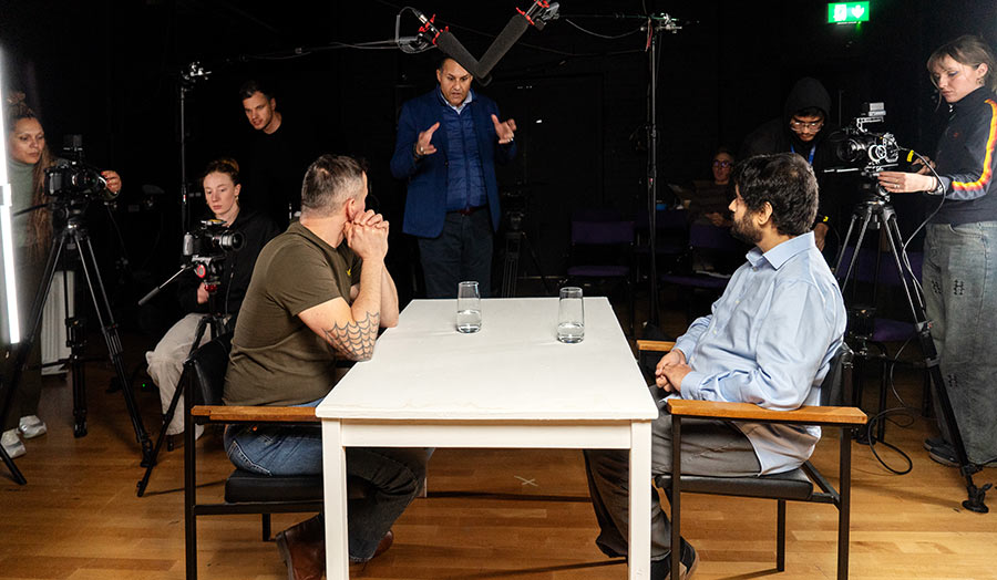 Two people sit at a table, watched by a film crew