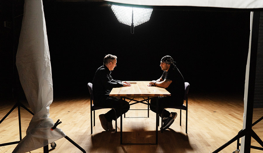 Two people sit at a brightly lit table in an otherwise dark film studio