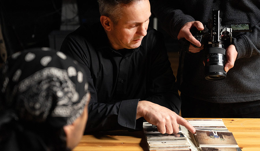 A person points at several photographs on a table, while someone points a handheld camera at the photos