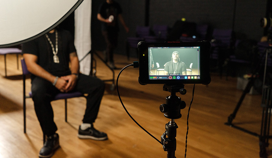 A person sits in a dark studio, partially obscured by lighting equipment, with a camera in front