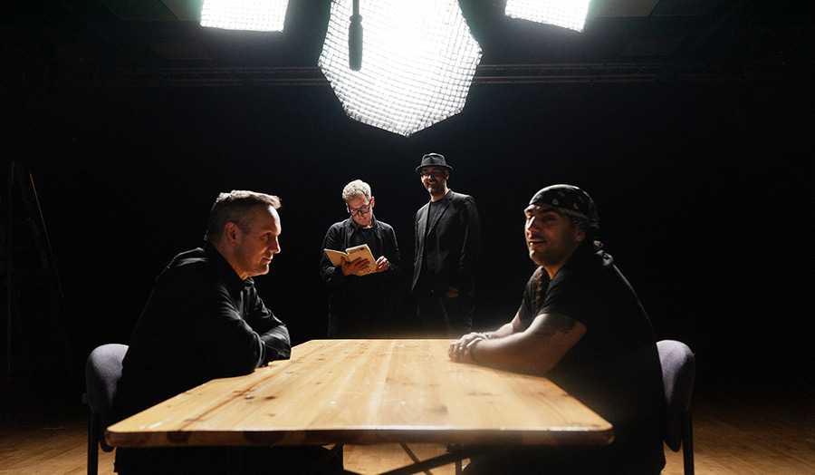 Two people sit at a table under bright studio lights, while others watch in darkness