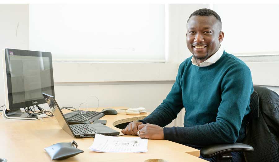 A computer science student smiling from his desk, working at a desktop screen and laptop